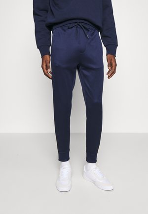 Homme portant un pantalon de survêtement bleu marine, un sweat bleu marine, des chaussettes blanches et des baskets blanches, debout devant un fond clair uni.