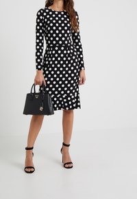 Svart långärmad klänning med vita prickar, figursydd midja och volangkant. En svart handväska med nyckelring och klackskor med ankelsnörning.