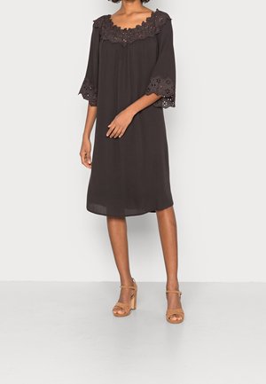 Kobieta w luźnej, czarnej sukience do kolan z wykończeniem z ażurowej koronki na dekolcie i rękawach, w połączeniu z brązowymi sandałami na wysokim obcasie.