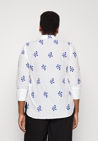 Bílá lněná košile s modrým květinovým vzorem, dlouhé rukávy a zakřivený lem. Obsahuje klasický límec a detaily s knoflíky.