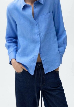 Osoba ubrana w jasno niebieską koszulę na guziki z kieszenią z przodu oraz ciemnoniebieskie spodnie ze sznurkiem, ręce w kieszeniach, na jednolitym tle.