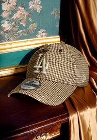 Bruine houndstooth pet met LA-logo, verstelbare band en zachte stoftextuur. Het label toont maat- en merkinformatie.
