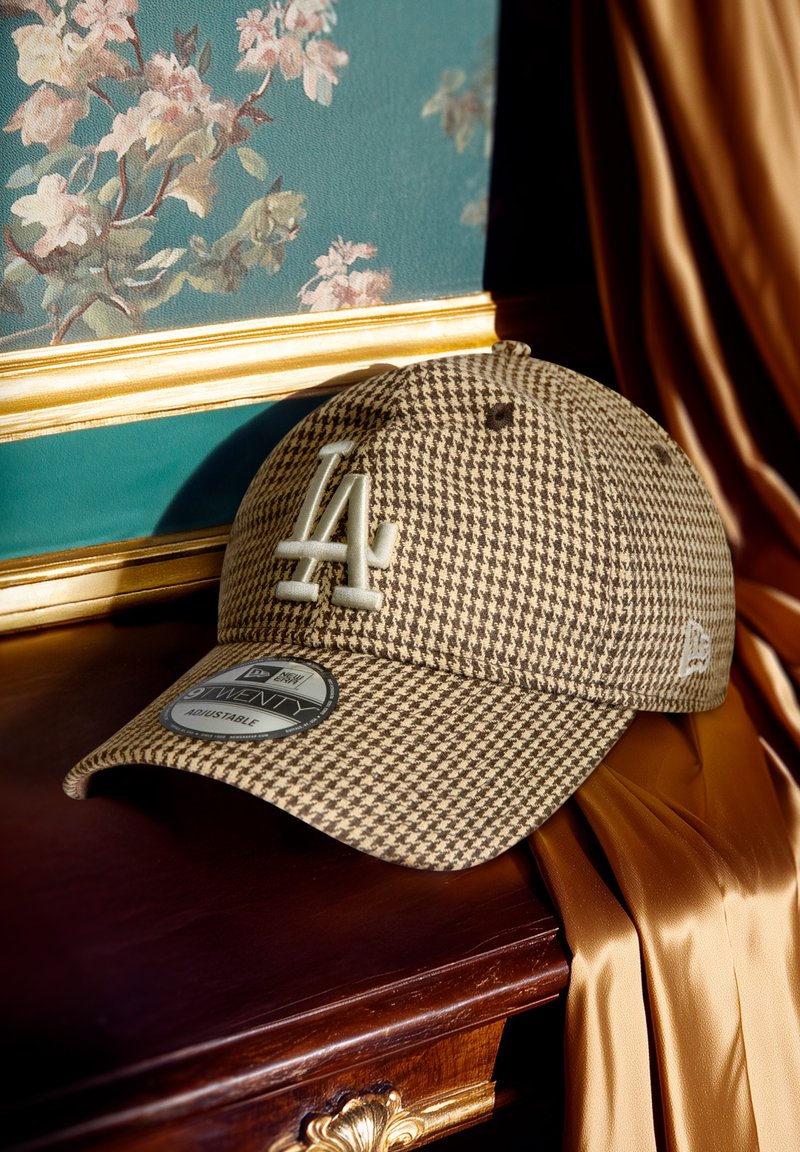 Bruine houndstooth pet met LA-logo, verstelbare band en zachte stoftextuur. Het label toont maat- en merkinformatie.