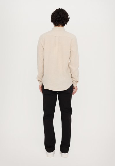 Chemise beige clair à manches longues avec un col à boutons, détail de patte au dos et manches retroussées, associée à un pantalon noir et des baskets blanches.