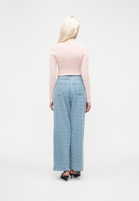 Γυναίκα με μακριά ξανθά μαλλιά, ντυμένη με ανοιχτό ροζ μακρυμάνικη μπλούζα, μπλε φαρδιά τζιν με σχέδια και μαύρα παπούτσια με τακούνι, στέκεται γυρισμένη προς τα πίσω.