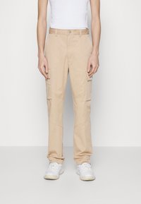 Beige cargobyxor med rak bendesign, utrustade med flera sidofickor och en mjuk textur, kombinerat med vita sneakers.
