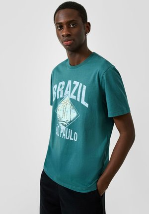 Junger Mann trägt ein türkisfarbenes T-Shirt mit dem Schriftzug "BRAZIL SÃO PAULO" und einer Palmen-Grafik, steht mit einer Hand in der Tasche vor einem grauen Hintergrund.