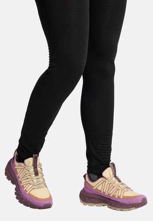 Beine, die schwarze strukturierte Leggings und beige Sneakers mit violetten und braunen Akzenten vor einem schlichten, hellen Hintergrund tragen.