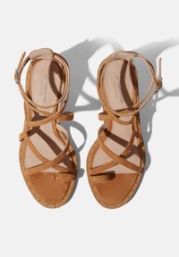 Narančaste suede sandale s više ukriženih traka, kopčom oko gležnja, ravnim potplatom i dekorativnim metalnim zakovicama duž ruba.