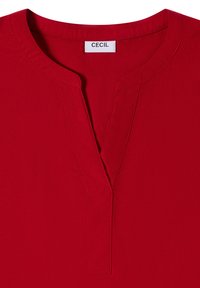 Blouse rouge avec un col en V et des coutures visibles autour du cou, avec une étiquette blanche portant le nom de la marque "CECIL."