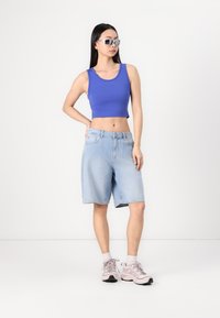 Blaues kurzes Tanktop, hellblaue Jeanskurzshorts und rosa Sportschuhe. Glatter Stoff, entspannter Schnitt, mittelkurze Shorts. Silberne Sonnenbrille.