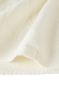 Bílý pletený deka s texturovaným kabelovým vzorem, měkkým pocitem a malou textilní etiketou s motivem králíčka na jednom okraji.