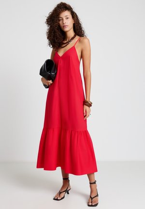Femme aux cheveux bouclés portant une robe midi rouge sans manches, des talons noirs à brides, tenant une pochette noire, debout devant un fond uni.