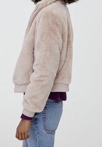 Faux fur bomberjakke i lyserød, med lynlås, ribbede manchetter og kant, båret over en lilla sweater med blå jeans.