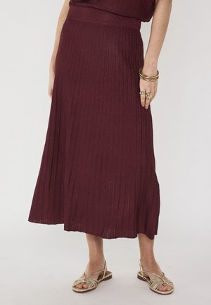 Femme portant une jupe midi côtelée rouge foncé avec des anneaux et bracelets en or, assortie à des sandales plates métalliques sur un fond uni.