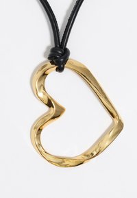 Colgante en forma de corazón de oro con un diseño ondulado, suspendido de un cordón de cuero negro, asegurado con un nudo. Superficie suave y brillante.