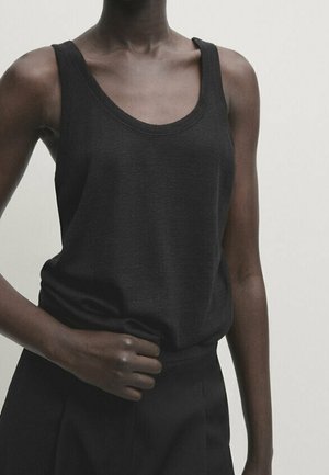 Ærmeløs sort tanktop båret af person med mørk hud, kombineret med højtaljede sorte bukser mod en ensartet lys baggrund.