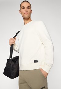 Sweatshirt crem cu guler rotund și manșete ribbate. Dispune de un etichetă în colțul din stânga jos. Modelul poartă o geantă neagră cu panou din plasă.