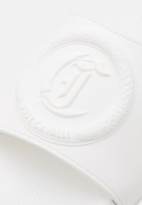 Sandale albe cu talpă plată, prezentând o curea texturată cu un logo circular embosat și denumirea brandului "Just Cavalli" pe margine.