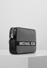 Svart och vit rektangulär handväska med strukturerat mönster, som har en framträdande vit "MICHAEL KORS"-logotyp och silverkedje-rem.