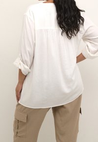 Blouse en lin blanc à coupe décontractée, avec un dos plissé et des manches retroussées. Associée à un pantalon cargo beige avec des poches latérales.