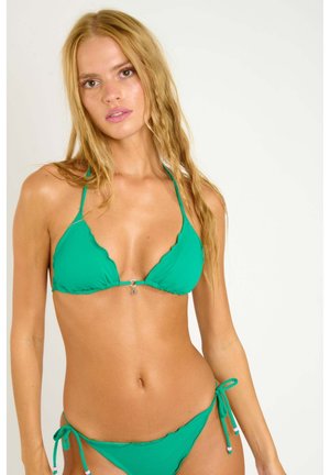 Femme aux longs cheveux blonds portant un bikini vert à bords festonnés, posant devant un fond blanc uni.