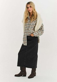 Košile na knoflíky se vzorem káro v zelených a krémových tónech přes šedou džínovou sukni. Hnědé semišové boty s kovovými detaily doplňují outfit.