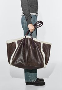 Person wearing jeans and sweater holding large dark brown shearling tote bag with white fur trim by its handles.