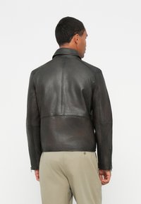 Veste en cuir noir avec un col, des coutures visibles et des zips sur le côté. Texture lisse, design ajusté et finition légèrement brillante.
