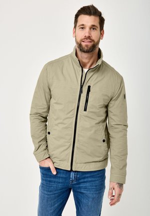 Bearded man wearing a light beige zip-up jacket with black zipper details and blue jeans, standing with one hand in pocket.