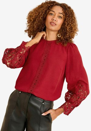 Frau mit lockigem Haar, die eine rote Bluse mit Spitzenärmeln und schwarze Lederhosen trägt, posiert mit einer Hand in der Tasche.