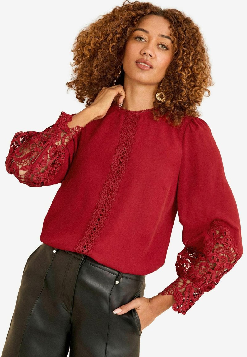 Donna con capelli ricci che indossa una blusa rossa con maniche in pizzo e pantaloni in pelle neri, mentre posa con una mano in tasca.