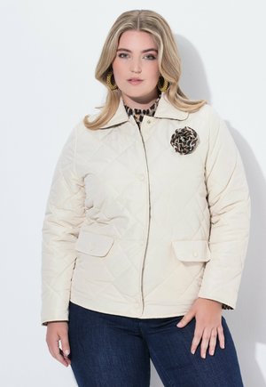 Femme aux cheveux blonds portant des boucles d'oreilles dorées, une veste matelassée beige avec un broche en imprimé léopard, et un jeans bleu foncé sur un fond blanc.