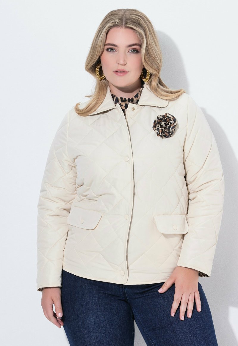 Kvinna med blont hår som bär guldörhängen i form av ringar, en beige, kviltad jacka med en leopardtryckt brosch och mörkblå jeans mot en vit bakgrund.