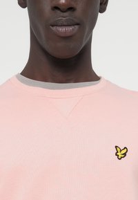 Ανοιχτό ροζ φούτερ με γκρι γιακά, διακοσμημένο με ένα μικρό κεντητό λογότυπο σε κίτρινο και μαύρο στην αριστερή πλευρά του στήθους. Μαλακή υφή, casual σχέδιο.