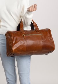 Borsa da viaggio in pelle marrone con due manici robusti, tasca con zip sulla parte frontale e forma arrotondata. Presenta cuciture visibili e accessori in metallo.