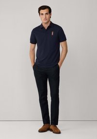 Marinblå polo-skorta med krage och korta ärmar, med en liten broderad design på vänster bröst. Åtföljd av mörka byxor och bruna skor.