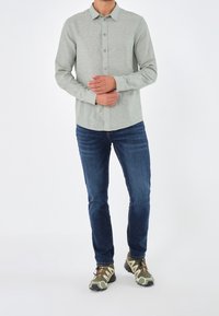 Uomo in camicia azzurro chiaro a maniche lunghe, jeans blu e scarpe da trekking verde mimetico in piedi con le mani giunte.
