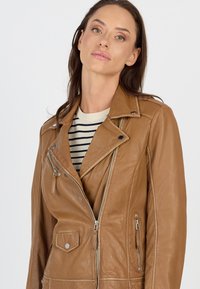 Braune Leder-Motorradjacke mit asymmetrischem Reißverschluss, Seitentaschen und einer Brusttasche. Über einem gestreiften Hemd getragen. Glatte Textur.