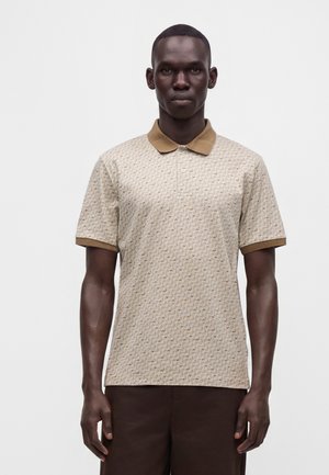 Homme debout portant un polo beige à motifs à manches courtes avec col et poignets marron, associé à un pantalon marron foncé.