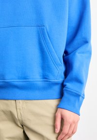 Sudadera de algodón azul con un bolsillo frontal, puños y dobladillo acanalados. Texture suave y diseño casual. Llevada con pantalones caqui.