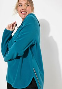 Pullover in pile verde acqua con maniche lunghe, aperture laterali e colletto con zip. Presenta una texture liscia e una vestibilità comoda.