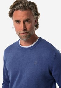 Stickad blå tröja med rund halsringning och broderad logotyp på bröstet. Materialet verkar mjukt och texturerat, lämpligt för avkopplande användning.