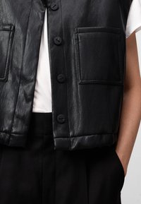 Gilet di pelle nera con una texture liscia, dotato di una patta frontale e due grandi tasche a toppa, abbinato a una camicia bianca sottostante.