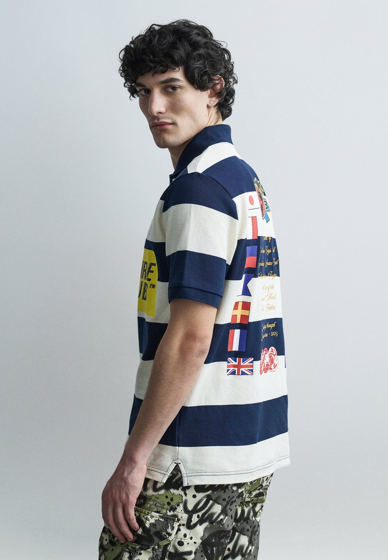 Νεαρός άνδρας με σγουρά μαλλιά που φοράει μπλε σκούρο και λευκό ριγέ πόλο με διάφορες εθνικές σημαίες και κείμενο στην πλάτη, και παντελόνι παραλλαγής.