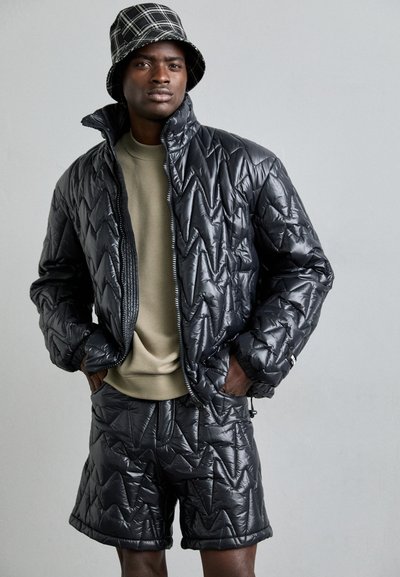 Schwarze Puffjacke mit einem gesteppten Zickzackmuster, kombiniert mit passenden Shorts. Hellgrauer Sweatshirt darunter. Schwarzer karierter Bucket Hat.