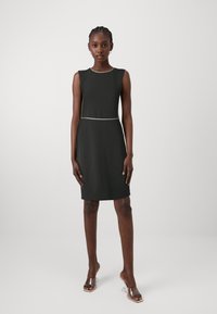 Vestido preto sem mangas com um design ajustado, apresentando uma linha branca em contraste na cintura. Textura suave, comprimento até ao joelho, estilo minimalista.