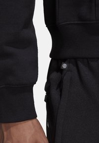 Svart sweatshirt med ribbstickade muddar visas på en arm, kombinerad med svarta joggers som har en liten logotypmärke och detalj med dragkedjeficka.