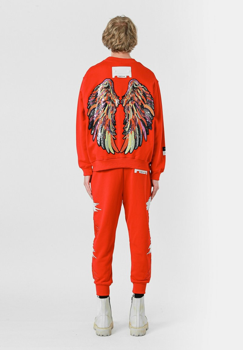 Sudadera roja con un diseño de alas multicolores en la espalda, combinada con pantalones a juego que presentan detalles desgastados. El calzado son botas blancas hasta el tobillo.
