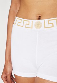 Shorts hipster bianchi in cotone con un motivo a chiave greca dorata e il simbolo della Medusa sull'elastico in vita. Texture liscia, forma aderente.
